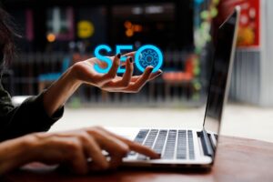 Person drückt eine Taste auf einem Laptop und hält in der anderen Hand den Schriftzug: SEO